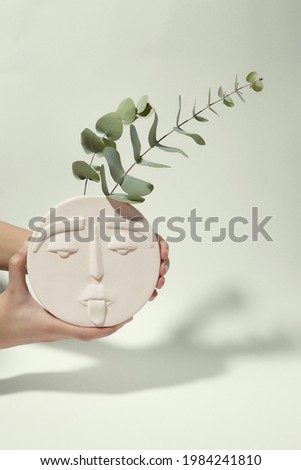 Cropped shot of female hands with white round ceramic vase shaped like human face. Two bluegum twigs are placed in modern creative flower vase.  