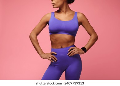Cropped shot of fit black woman in purple sportswear standing with her hands on hips over pink studio background. Unrecognizable fitness model posing with tracker smartwatch
