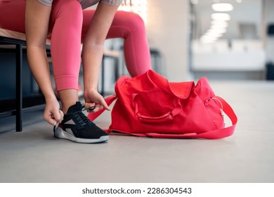 Cropped shot of fit sports woman in sportswear with gym bag wearing pink yoga pants and sneakers sitting on bench, getting ready for exercise session, tying her shoelaces in locker room at gym. - Powered by Shutterstock