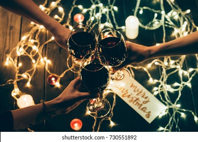 Cropped Shot Of Four People Clinking Glasses With Red Wine Over Wooden Table With Fairylights And Merry Christmas Card