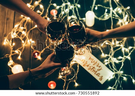 Cropped shot of four people clinking glasses with red wine over wooden table with fairylights and merry christmas card
