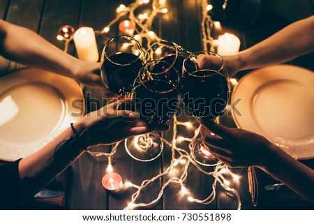 Cropped shot of four people clinking glasses with red wine over served table with fairylights decorations