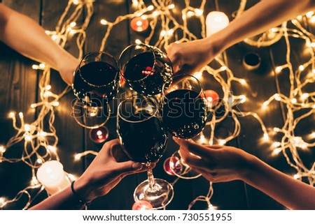 Cropped shot of four people clinking glasses with red wine over wooden table with fairylights decorations
