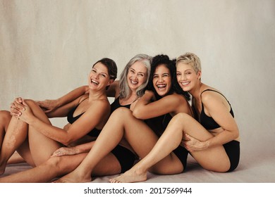 Cropped Shot Of Four Women Embracing Their Natural Bodies. Women Of All Ages Wearing Black Underwear And Smiling Cheerfully. Happy Women Sitting Together Against A Studio Background.