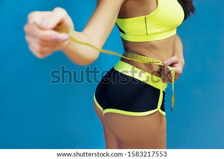 Cropped shot of girl in sportswear measuring herself with metric tape at blue background. Girl in sport top and short checking her waist size with tape. Measure of waist circumference by fit girl.
