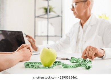 Cropped shot of green apple and measuring tape on dietitian desk. Healthy balanced diet eating concept. Caucasian elderly senior female doctor nutritionist female patient good nutrition habits.