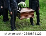 Cropped shot of group of intercultural men in black attire holding by handles of wooden coffin with white flowers on top of closed lid