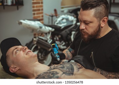 Cropped Shot Of A Handsome Young Man Getting Tattooed On Chest By Professional Tattoo Artist. Bearded Male Tattooist Working At His Studio, Making Tatoo On Chest Of His Client