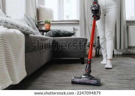 cropped shot of housewife in white jeans using cordless handheld vacuum cleaner in living room with sofa and pillows, household chores concept