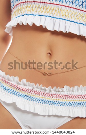 Cropped shot of lady's tummy, wearing golden waist chain, adorned with an insertion in view of infinity sign. The girl with tanned skin is wearing white swimsuit with colorful ornament and ruffles.