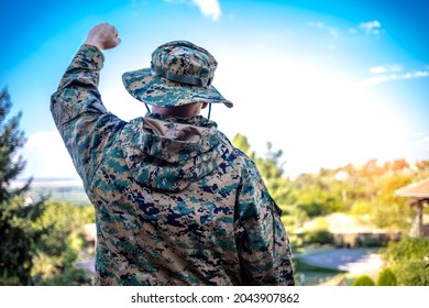 Cropped shot of a male army soldier saluting against blue sky. Soldier in the mountain forest, celebrating freedom. Rear view shot of a young soldier standing at the nature and saluting, looking away.