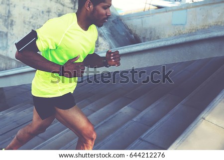 Cropped shot male dark-skinned athlete running up a flight of stairs with speed, sporty young man in fluorescent t-shirt training or working out outdoors while jogging up the steps, filtered image