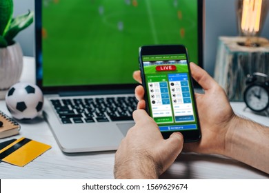 Cropped shot of male fan making bets using gambling mobile application on his phone. Man watching football play online broadcast on his laptop waiting for winning results.