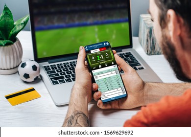 Cropped shot of male fan making bets using gambling mobile application on his phone. Handsome bearded man watching football play online broadcast on his laptop waiting for winning results. - Shutterstock ID 1569629692
