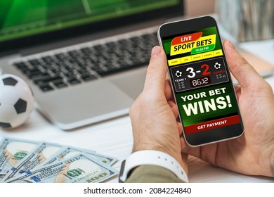 Cropped shot of male hands holding smartphone with bookmaker's mobile application interface design template. Man making bets while watching soccer play live broadcast online on his laptop. - Shutterstock ID 2084224840