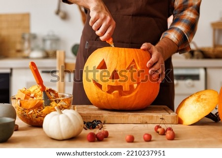 Cropped shot of  man in apron standing in kitchen and carving large orange pumpkin for Halloween party while  making scary face on jack-o-lantern with knife to set the mood for trick-or-treaters 