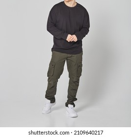 Cropped Shot Of Man In Black Blank Long Sleeved And Green Cargo Pants. Standing On Gray Background. Mockup For Print Or Design Template. Basic Clothing Line No Logo