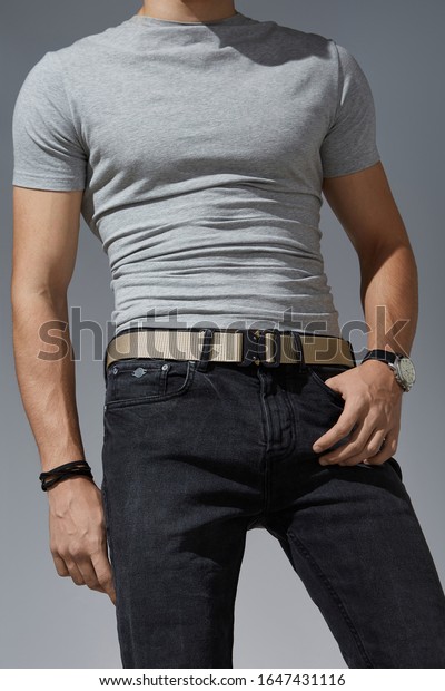 Cropped shot of a man in\
black jeans and a gray t-shirt on the gray background. A beige\
canvas belt is fitted with a black metal male buckle and a black\
belt loop.