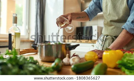 Cropped shot of man, chef cook pouring a glass of white wine into the pan with chopped vegetables while preparing a meal in the kitchen. Cooking at home, Italian cuisine. Selective focus. Web Banner