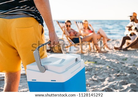 cropped shot of man holding beach cooler while friends resting on sand behind