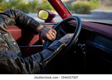 Cropped Shot Of Man In Leather Jacket With His Hands On Steering Wheel Of An Old Red Muscle Car. Stylish Guy Wearing Hipster Accessories Driving Alone. Close Up, Copy Space, Background.