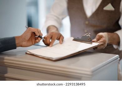 Cropped Shot Of Man Signing Papers Checking In At Hotel Reception Indoors. Male Staff Showing To Sign Documents Meeting Guest, Closeup Of Hands. Travel Accomodation Concept. Selective Focus