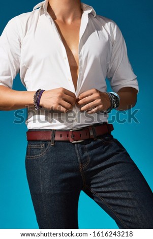 Cropped shot of a man in a white shirt, navy jeans and a russet leather belt with a metal buckle and a loop with leather inserts. The man with cord bracelets and wrist watch is unbuttoning his shirt. 