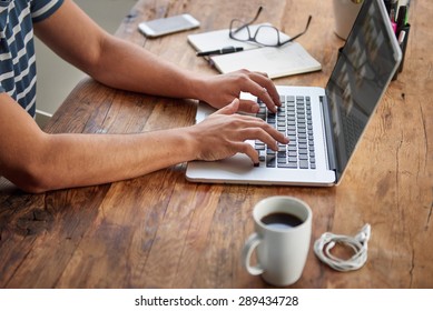 Cropped shot of a man's hands typing on a laptop that is on a wooden desk with a mug of coffee alongside