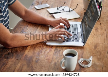 Cropped shot of a man's hands typing on a laptop that is on a wooden desk with a mug of coffee alongside