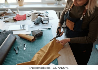 Cropped shot of mature female leatherworker cutting piece of suede or leather with scissors while working over new creative apparel - Powered by Shutterstock