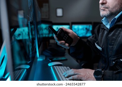 Cropped shot of mature male security guard with walkie-talkie in hand typing on computer keyboard and looking at screen by workplace