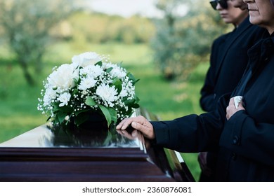 Cropped shot of mature woman and her family in mourning attire standing in front of coffin with closed lid with fresh white chrysanthemums