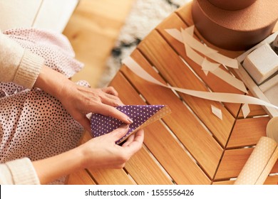 Cropped Shot Of Mature Woman Sitting In Living Room On Couch Packing Christmas Gift, Making Origami Wrap. Top View Of Senior Woman's Hands Holding Paper, Using Scissors And Ribbon, Wrapping Presents