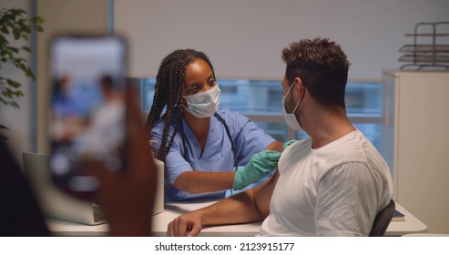 Cropped Shot Of Medical Staff Taking Photo Of Patient Getting Covid-19 Vaccine. African-American Female Nurse Inject Coronavirus Vaccine Into Man Hand In Hospital