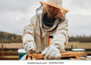 Cropped shot of middle aged male beekeeper in protective bee suit against grey autumn sky working with hive frame during honey harvesting at apiary farm, copy space - Powered by Shutterstock