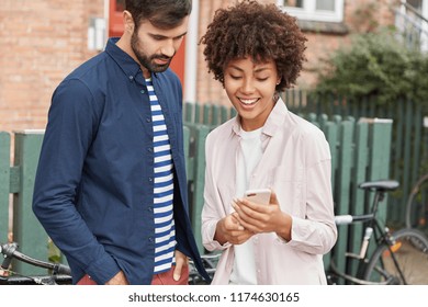 Cropped Shot Of Mixed Race Friends Have Rest After Riding Bicycles, Stand Closely, Looks Photos On Cell Phone, Pose Outdoor. African American Woman And Caucasian Guy Watch Video On Gadget Outside