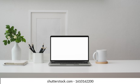 Cropped shot of modern workspace with blank screen laptop, frame, pencils, coffee cup and vase on white table with white wall background     - Shutterstock ID 1610162200