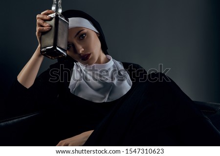 Cropped shot of a nun, sitting on a black chair. She's wearing dark nun's clothing. The nun is holding bottle of whiskey in right hand at her forehead. She is looking at camera. 