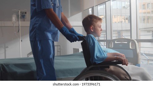 Cropped Shot Of Nurse Pushing Sick Boy In Wheelchair In Hospital Ward. Medical Worker Transporting Disabled Kid Patient In Wheelchair To Window In Hospital Room