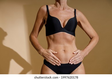 Cropped Shot Of Perfect Fit Body Of Mature Woman In Black Underwear Holding Hands On Her Stomach While Posing Half Naked, Standing Isolated Over Beige Background