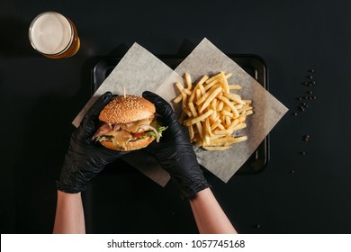 Cropped Shot Of Person In Gloves Holding Tasty Burger Above Tray With French Fries And Glass Of Beer On Black