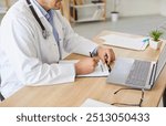 Cropped shot of physician or cardiologist in lab coat sitting at desk in front of laptop. Male doctor filling out report or writing prescription after patient medical health examination in clinic.