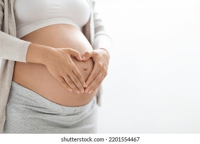 Cropped shot of pregnant woman in homewear holding hands in heart-shaped symbol gesture on big belly, unrecognizable lady waiting for baby, white studio background, copy space, closeup - Powered by Shutterstock