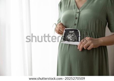 Cropped shot of pregnant woman showing unborn baby sonogram image at camera. Expecting mom holding ultrasound scan picture at big belly. Medical examination, prenatal care, pregnancy concept