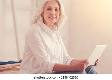 Cropped shot of a pretty retired lady sitting on the edge of her bed and smiling while looking into the camera with a computer on her knees.