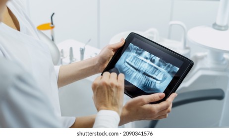 Cropped shot of professional dentist showing jaws and teeth x-rays to her patient, using a digital tablet technology, discussing a treatment plan while they standing in stomatology clinic, web banner - Powered by Shutterstock