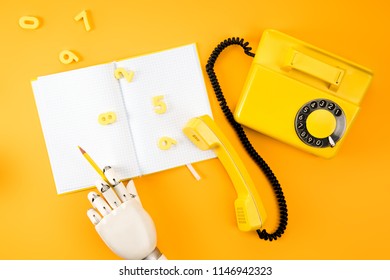 Cropped Shot Of Robotic Hand Writing In Blank Notebook On Yellow Tabletop With Vintage Phone And Math Numbers