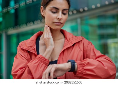 Cropped shot of serious woman checks pulse on neck monitors fitness activity has quick heart rate wears red jacket poses outdoors against blurred background controls her health. Devices for sport