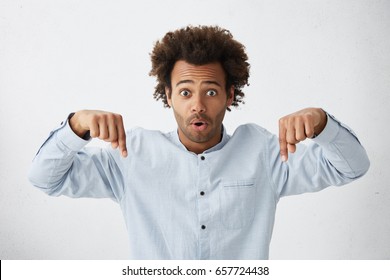 Cropped Shot Of Shocked Man With Curly Bushy Hair And Dark Eyes Wearing Formal Shirt Pointing Down With Fingers Looking With Opened Mouth At Camera. People, Emotions, Feelings, Body Language Concept
