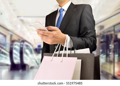 Cropped Shot Of Shopper Man Holding The Shopping Bags An Using The Smart Phone At The Center Mall. Concept Of Shopaholic Man.